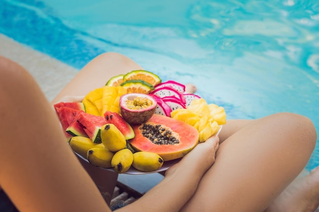 Jonge vrouw ontspannen en eten fruitschaal bij het zwembad van het hotel. Exotisch zomerdieet. Foto van benen met gezond voedsel bij het zwembad, bovenaanzicht van bovenaf. Tropisch strand levensstijl.