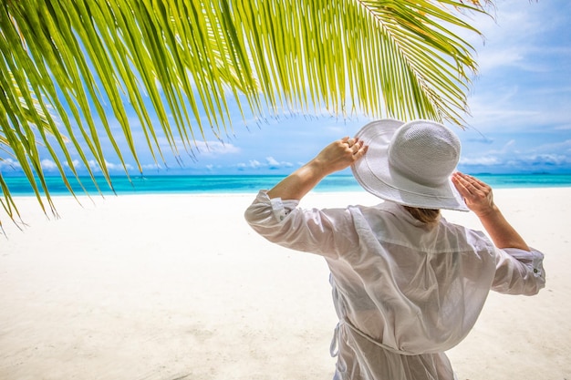 Jonge vrouw ontspant op een tropisch strand, witte jurk witte hoed onder palmblad. Luxe levensstijl