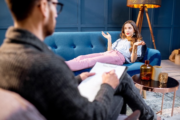 Jonge vrouw op bezoek bij mannelijke psycholoog liggend op de comfortabele bank tijdens psychologische sessie in het luxe blauwe kantoorinterieur