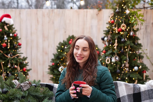 Jonge vrouw op de achtergrond van de kerstboom met een kopje koffie buiten Achtertuin decoratie nieuwjaar