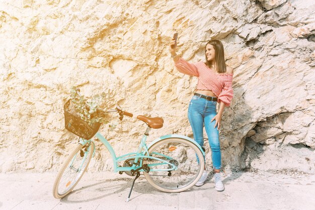 Jonge vrouw op een fiets neemt een selfie met haar mobiele telefoon naast een stenen muur