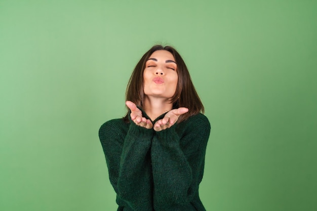 Jonge vrouw op groen in een gezellige grote gebreide trui schattig stuurt een kus naar de camera