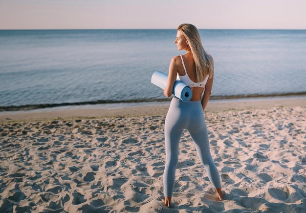 Jonge vrouw op het strand in een trainingspak met een yogamat