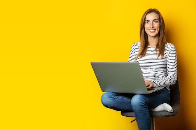 Jonge vrouw op stoel die bij laptop op gele achtergrond werkt.