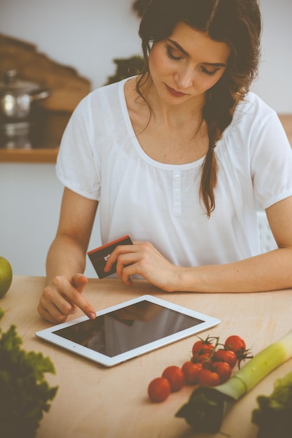 Jonge vrouw op zoek naar een nieuw recept voor koken in een keuken. Huisvrouw maakt online winkelen via tabletcomputer en creditcard.