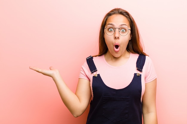 Jonge vrouw op zoek verrast en geschokt, met kaak gevallen met een object met een open hand aan de zijkant tegen roze muur