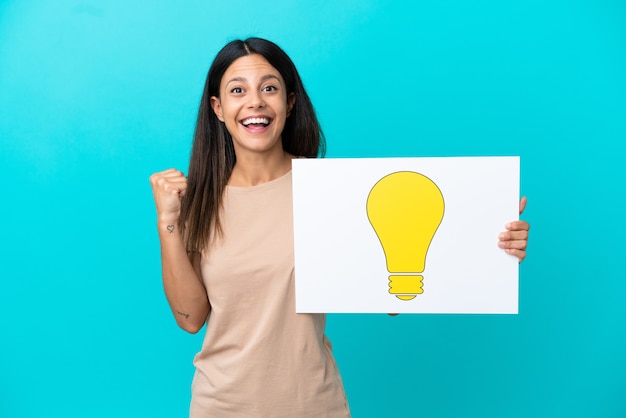 Jonge vrouw over geïsoleerde achtergrond die een plakkaat met bolpictogram houdt en een overwinning viert