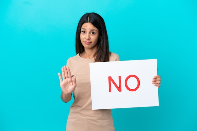 Jonge vrouw over geïsoleerde achtergrond die een plakkaat met tekst NEE houdt en stopbord doet