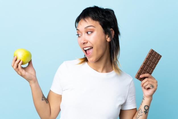 Jonge vrouw over geïsoleerde blauw met een chocoladetablet in de ene hand en een appel in de andere