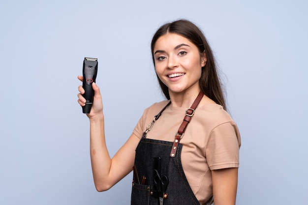 Jonge vrouw over geïsoleerde blauw met kapper of kapper jurk en haar snijmachine te houden