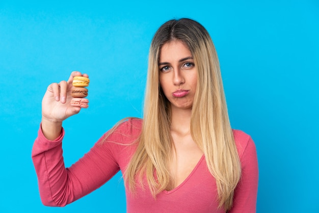 Jonge vrouw over geïsoleerde blauwe muur die kleurrijke Franse macarons en met droevige uitdrukking houdt