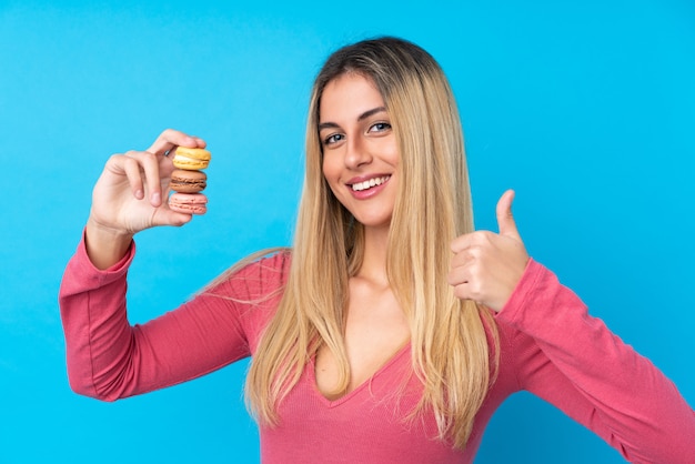 Jonge vrouw over geïsoleerde blauwe muur die kleurrijke Franse macarons met omhoog duimen houdt