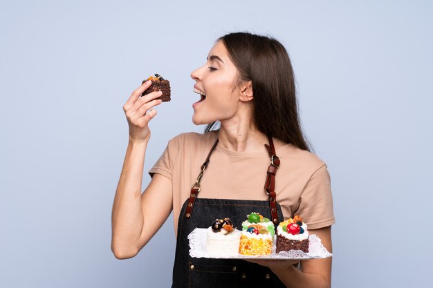 Jonge vrouw over geïsoleerde blauwe muur die minicakes houden en het eten