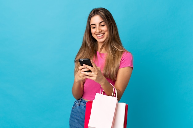 Jonge vrouw over geïsoleerde blauwe muur met boodschappentassen en het schrijven van een bericht met haar mobiele telefoon naar een vriend