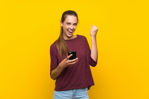 Jonge vrouw over geïsoleerde gele achtergrond met telefoon in overwinningspositie