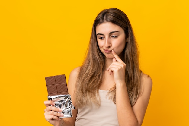 Jonge vrouw over geïsoleerde gele muur die een chocoladetablet neemt en twijfels heeft