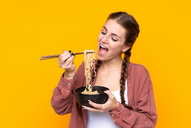 Jonge vrouw over geïsoleerde gele muur die een kom van noedels met eetstokjes houdt en het eet