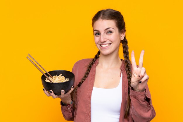 Jonge vrouw over geïsoleerde gele muur die en overwinningsteken glimlachen tonen terwijl het houden van een kom van noedels met eetstokjes