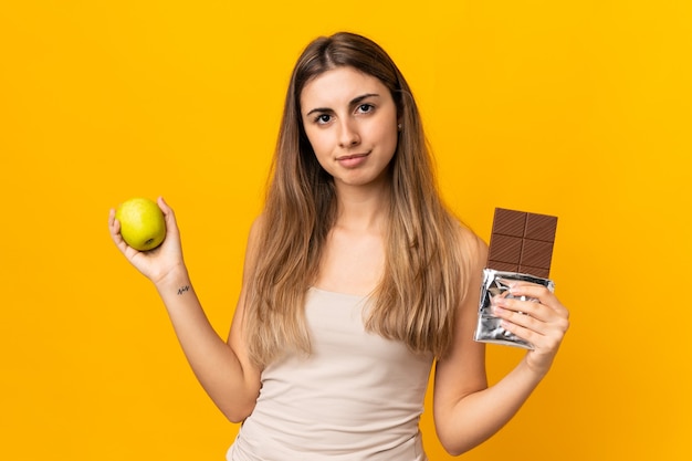 Jonge vrouw over geïsoleerde gele muur die twijfels heeft tijdens het nemen van een chocoladetablet in de ene hand en een appel in de andere