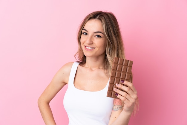 Jonge vrouw over geïsoleerde roze muur die een chocoladetablet nemen en gelukkig