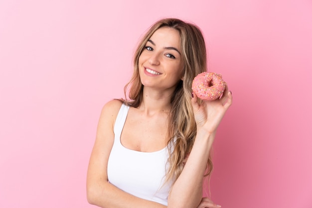 Jonge vrouw over geïsoleerde roze muur die een doughnut houdt en gelukkig