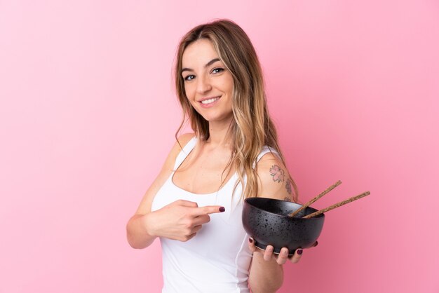 Jonge vrouw over geïsoleerde roze muur en het richten terwijl het houden van een kom noedels met eetstokjes