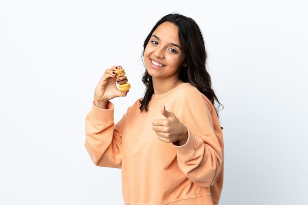 Jonge vrouw over geïsoleerde witte achtergrond die kleurrijke Franse macarons met omhoog duimen houdt