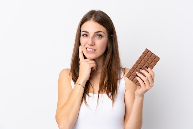 Jonge vrouw over geïsoleerde witte muur die een chocoladetablet neemt en twijfels heeft