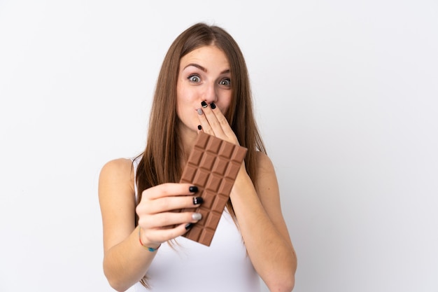 Jonge vrouw over geïsoleerde witte muur die een chocoladetablet neemt en verrast