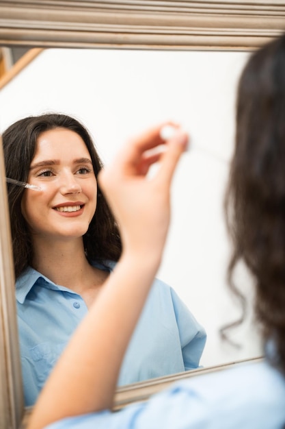 Foto jonge vrouw past vochtgevend serum op haar huid gezicht kijkt in de spiegel meisje met hyaluronzuur of serum pipette in handen close-up