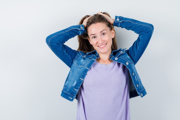 Jonge vrouw poseren met handen op het hoofd in t-shirt, jas en er vrolijk uitzien. vooraanzicht.