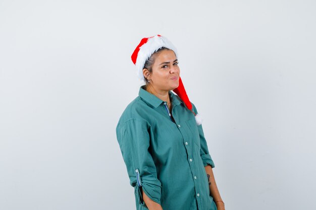 Jonge vrouw poseren terwijl ze in shirt, kerstmuts staat en er zelfverzekerd uitziet, vooraanzicht.