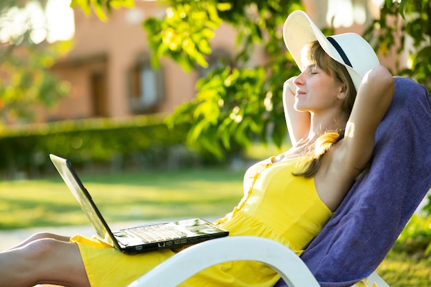 Jonge vrouw rusten in groene tuin op een stoel na het werk op laptop aangesloten op draadloos internet in zomerpark. Zakendoen en op afstand studeren tijdens quarantaine op vakanties concept.