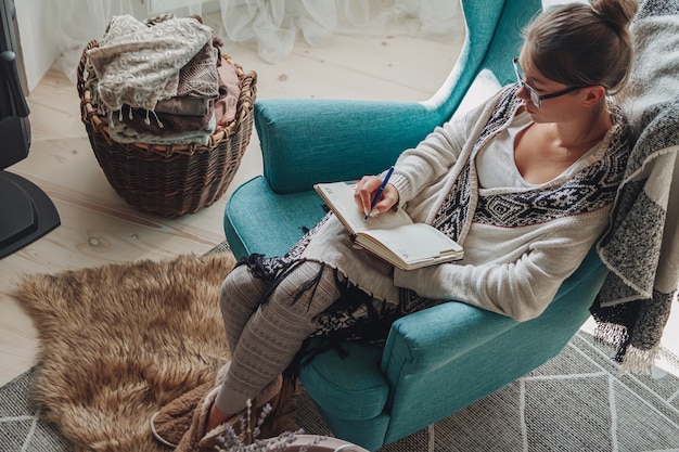 Jonge vrouw schrijft in een notitieboekje zittend in een gezellige fauteuil bij de open haard