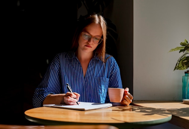 Jonge vrouw schrijft plannen in notitieboekje zittend aan houten bureau in café met koffiekopje en zonlicht