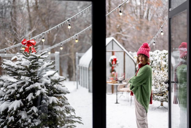 Jonge vrouw siert kerstboom met feestelijke ballen terwijl ze op het terras staat in een besneeuwde achtertuin Meisje dat zich voorbereidt op een wintervakantie