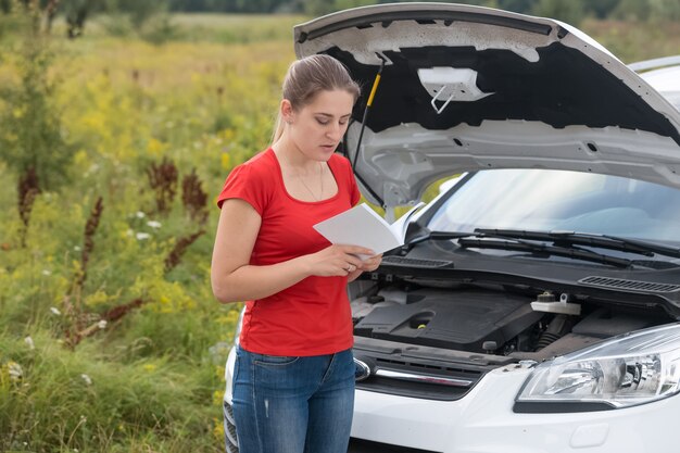 Jonge vrouw staat bij kapotte auto in de weide en leest de gebruikershandleiding reading