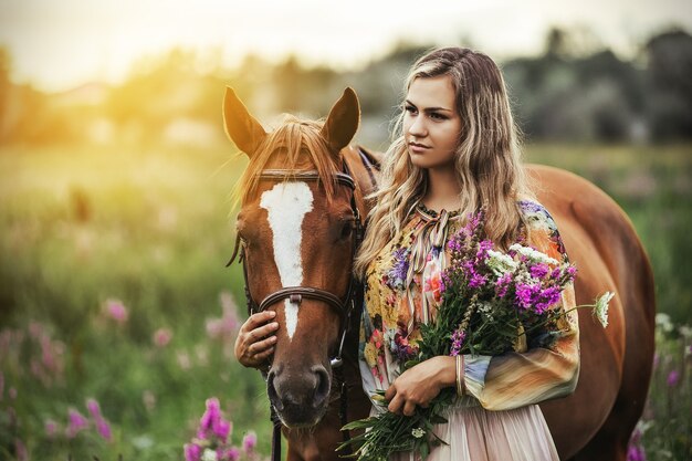 Jonge vrouw staat in de buurt van een paard in een zomerjurk op een weide