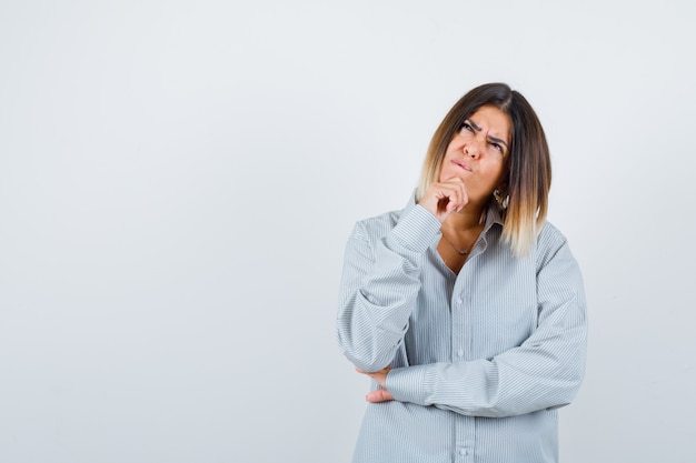 Jonge vrouw staat in denkende pose in oversized shirt en kijkt peinzend. vooraanzicht.