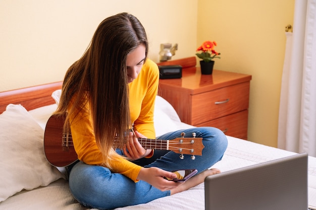 Jonge vrouw studeerde met ukelele bovenop bed met laptop