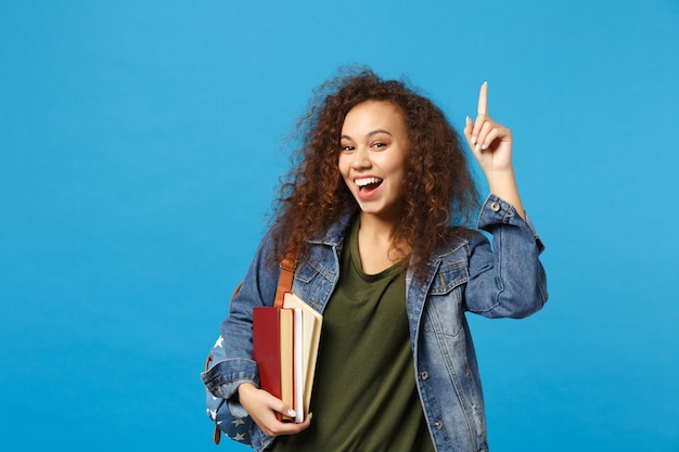 Jonge vrouw student in denim kleding en rugzak houdt boeken geïsoleerd op blauwe muur