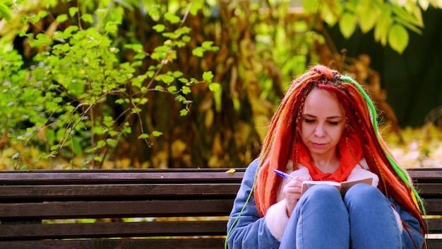 Jonge vrouw student met lang haar in vrijetijdskleding zittend op een houten bankje en het schrijven van notities in notebook