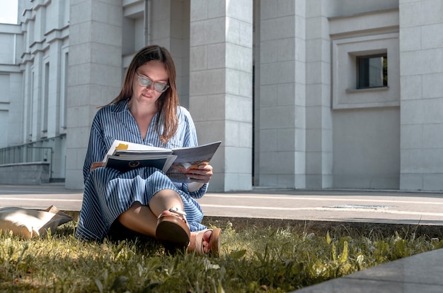 Jonge vrouw student zittend op het gras in de buurt van universiteit en leren schrijven en lezen met academische...