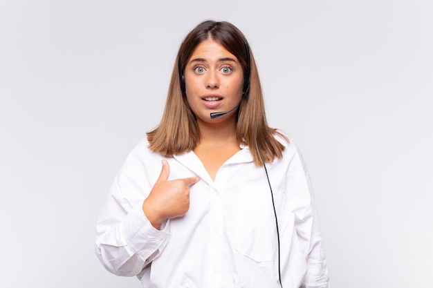 Jonge vrouw telemarketeer voelt zich gelukkig, verrast en trots, wijzend naar zichzelf met een opgewonden, verbaasde blik