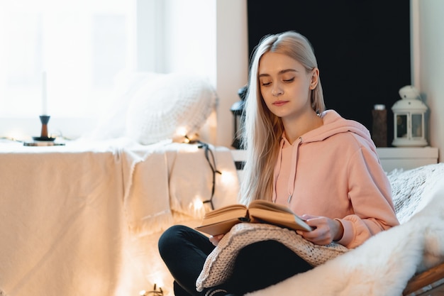 Jonge vrouw thuis koelen in een comfortabele ronde fauteuil bij het raam. meisje ontspannen, lezen en dromen van boek in de woonkamer van de loft.