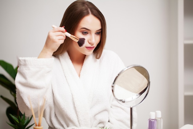 Jonge vrouw thuis past make-up toe op het gezicht in de badkamer voor een spiegel.