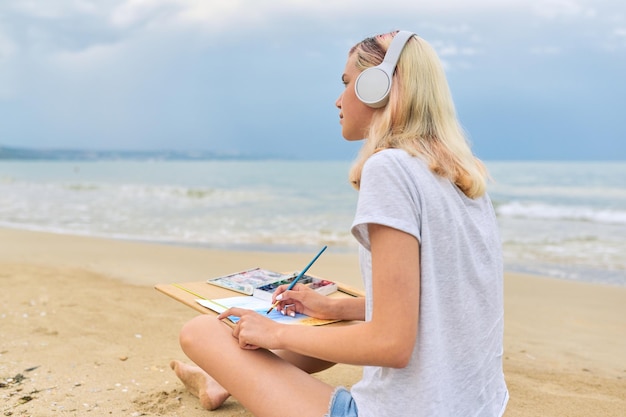 Jonge vrouw tiener in koptelefoon tekenen schets met aquarellen zittend op zee strand