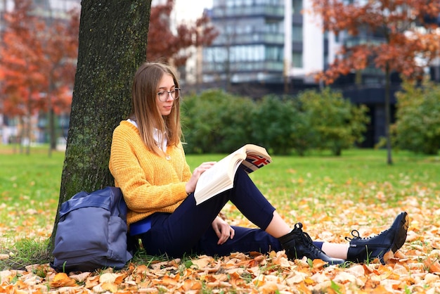 Jonge vrouw, tienermeisje, zittend op een grond in het gouden herfstpark op bladeren met rugzak erin