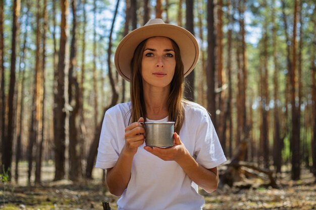 Jonge vrouw toerist in hoed en t-shirt drinkt thee of water tijdens een stop in het bos.