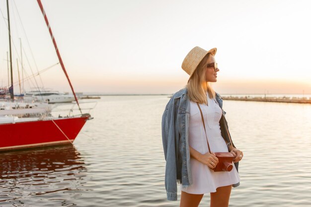 Jonge vrouw toerist met een camera op het strand bij zonsopgang gekleed in een hoed, witte jurk en zonnebril